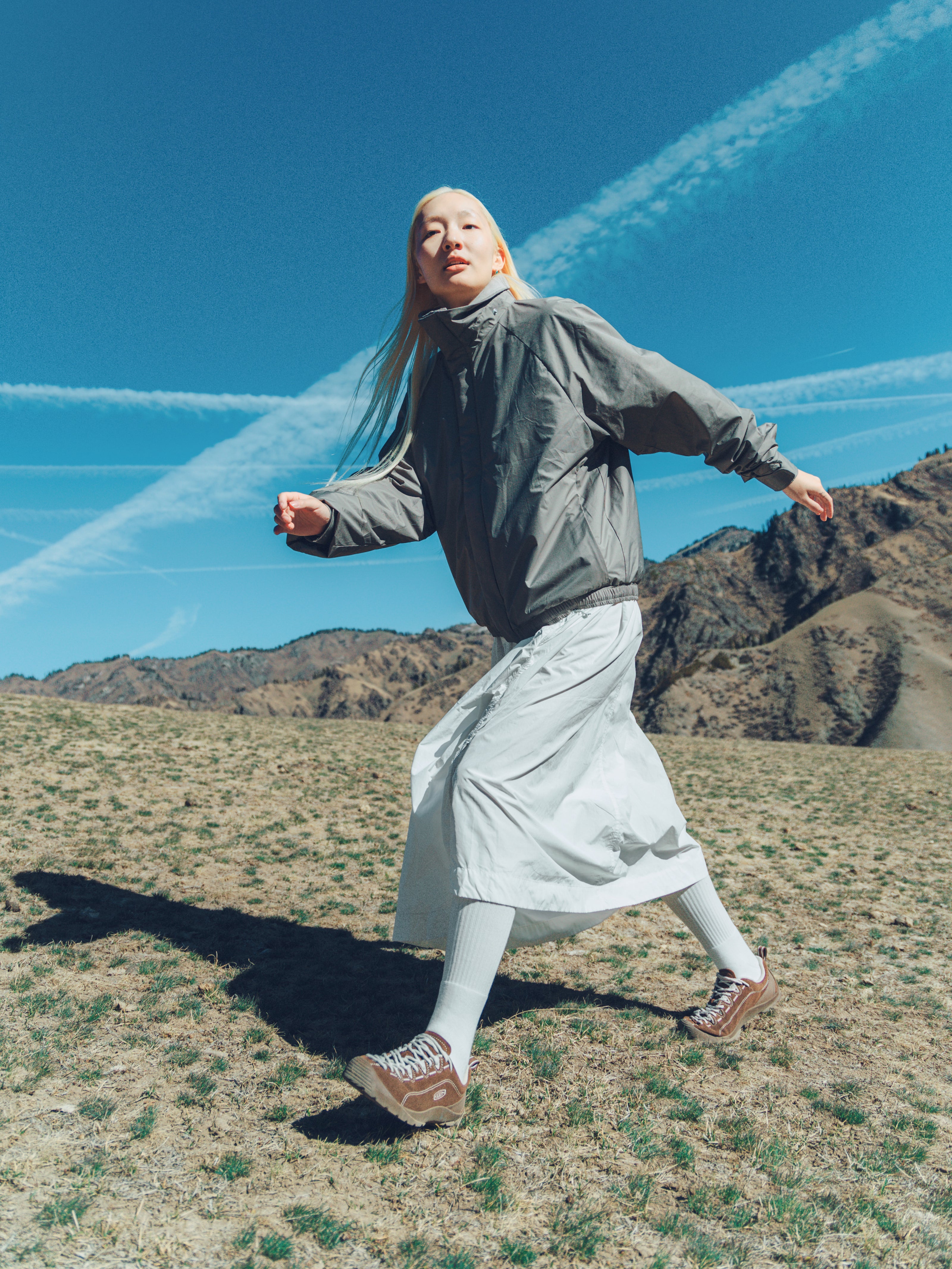 A woman wearing a gray lightweight jacket, white midi skirt, white tights, and brown sneakers is running across a dry grassy field with mountains and a blue sky with contrails in the background.