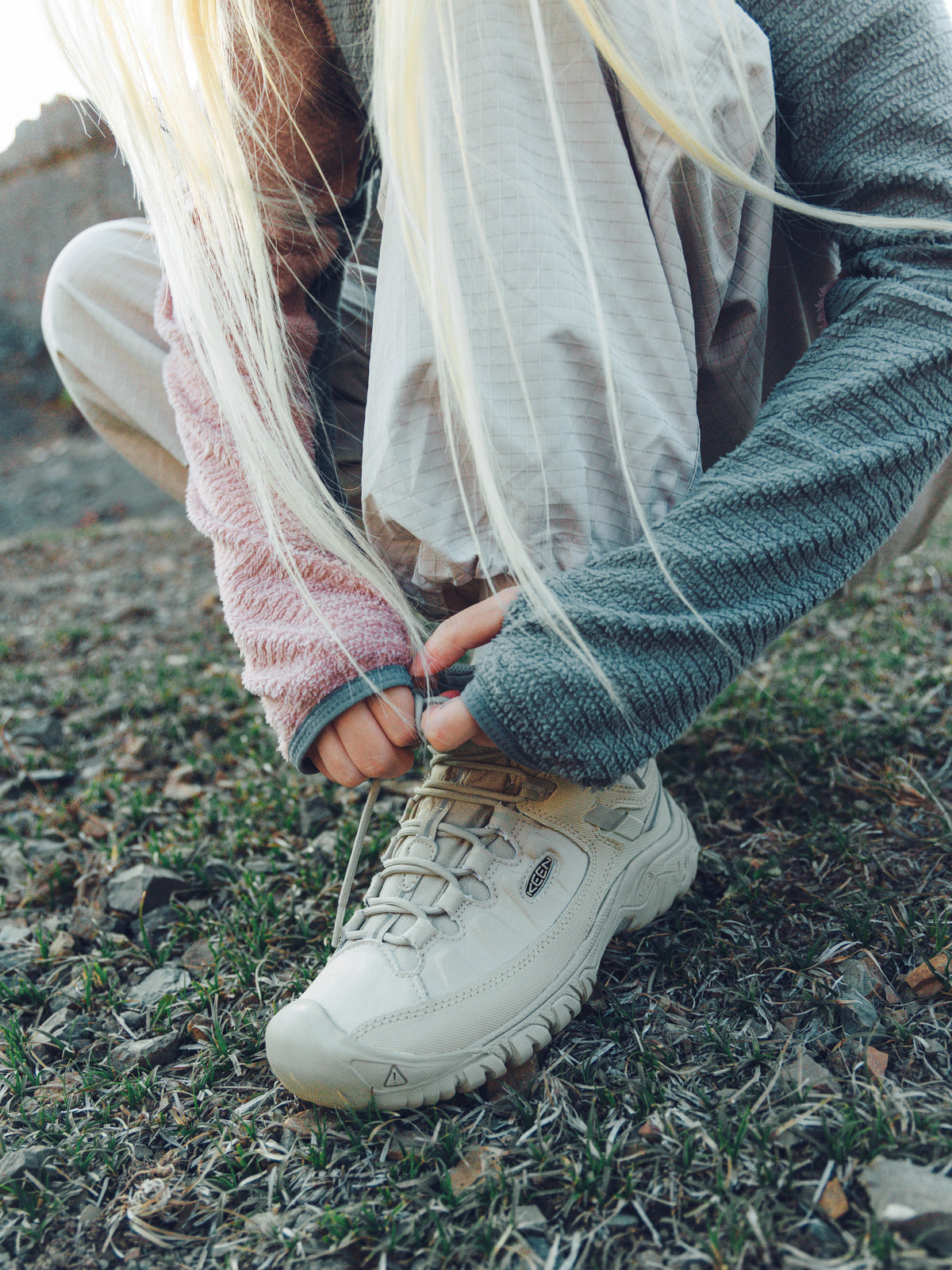 A close-up outdoor shot of a person crouching on grass and dirt while tying the laces of a beige