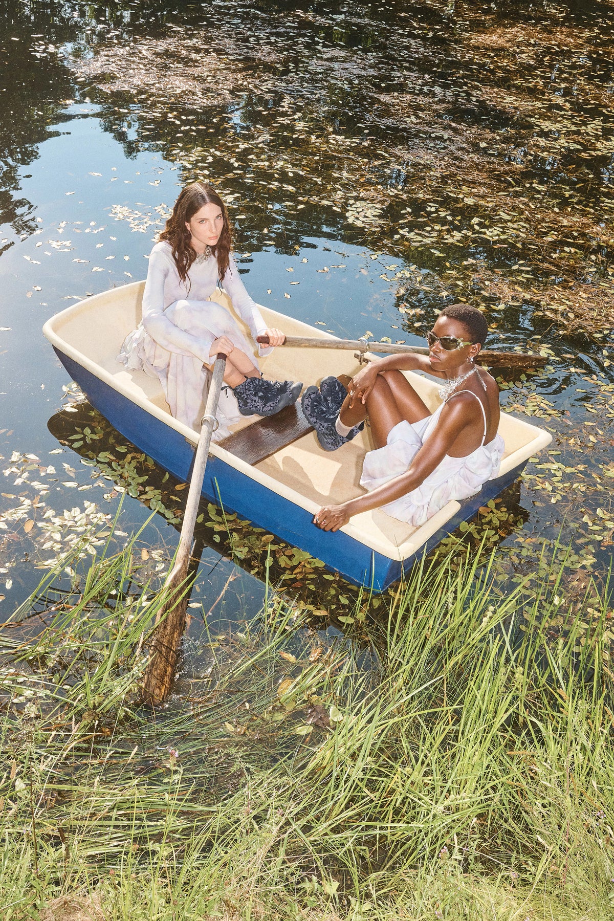 Two models in a blue and cream rowboat on a lake, styled in white dresses