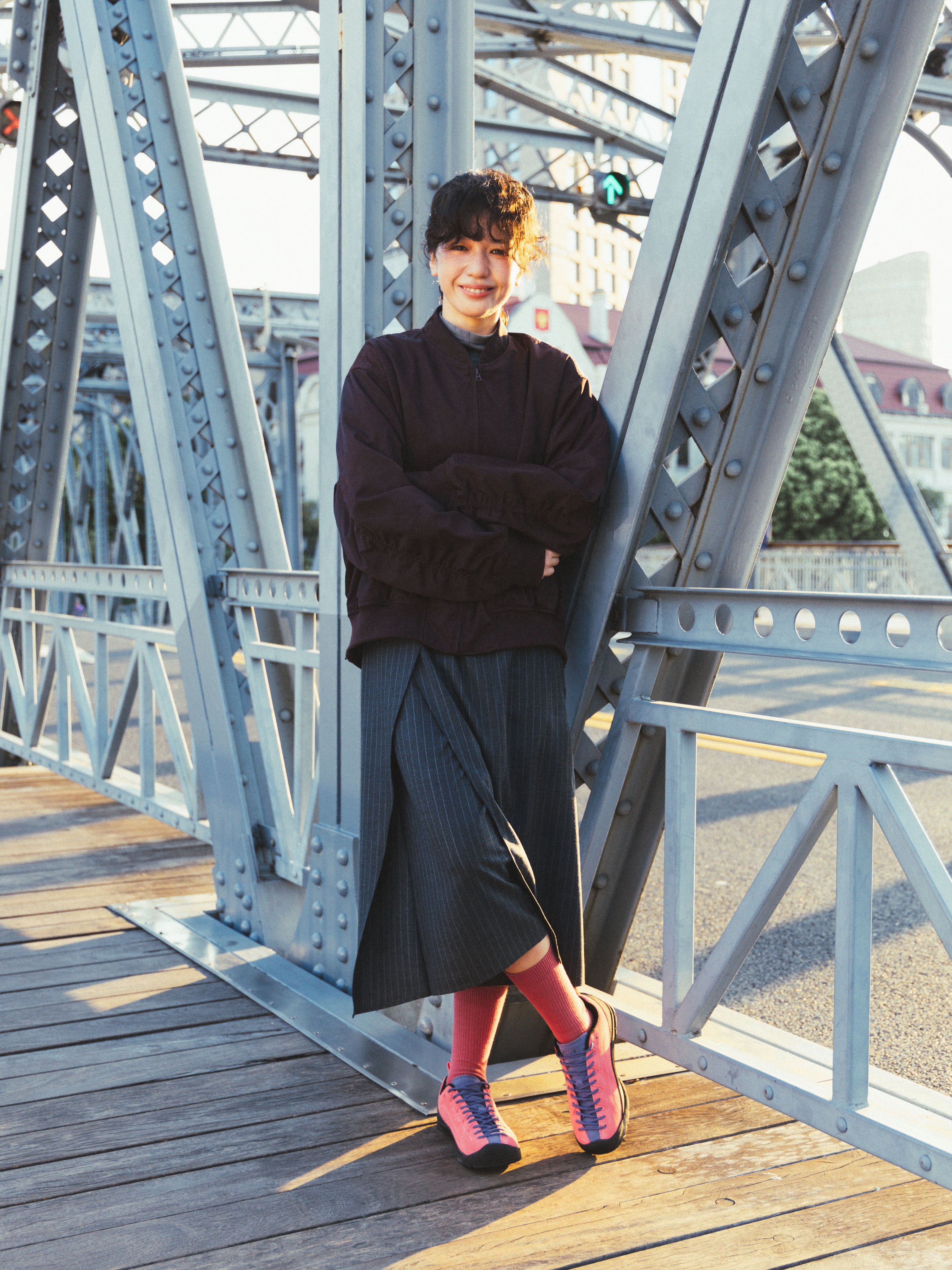 A woman stood on a bridge, wearing a dark brown long-sleeve top, a dark pleated skirt, pink knee-high socks, and pink-and-blue sneakers.