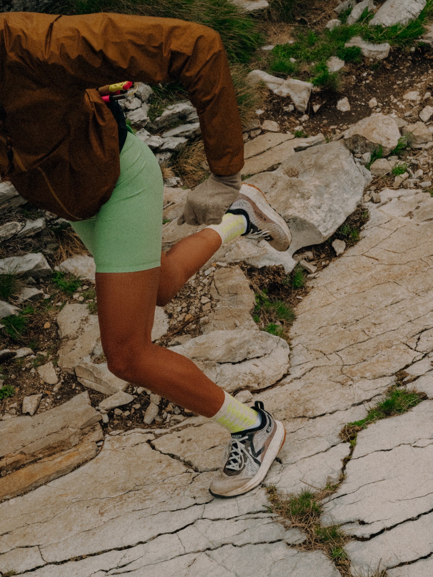 Person in green shorts and brown jacket jumping on a rocky path