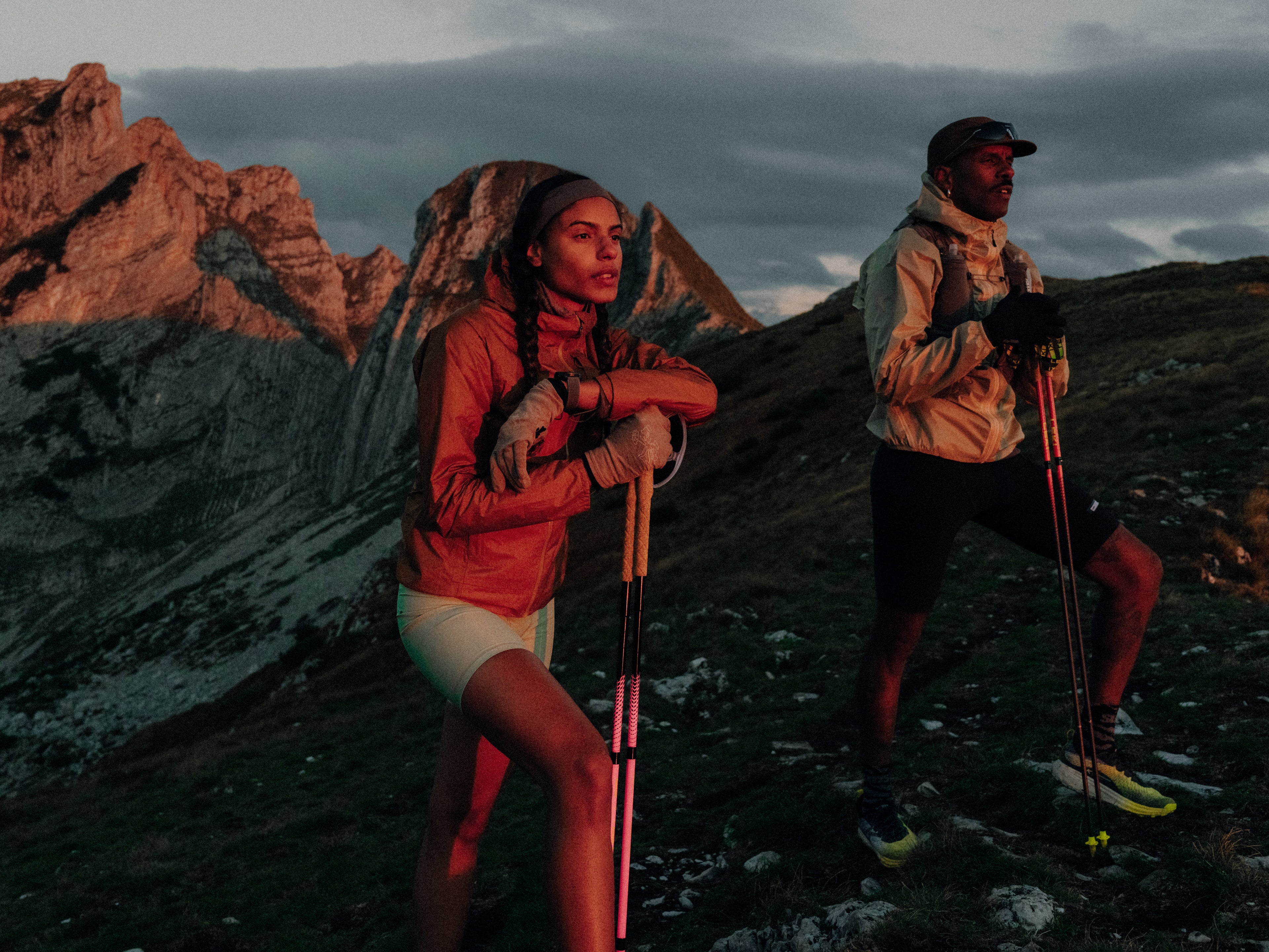 Two hikers with backpacks and hiking poles standing on a mountain trail with rocky cliffs and overcast sky.