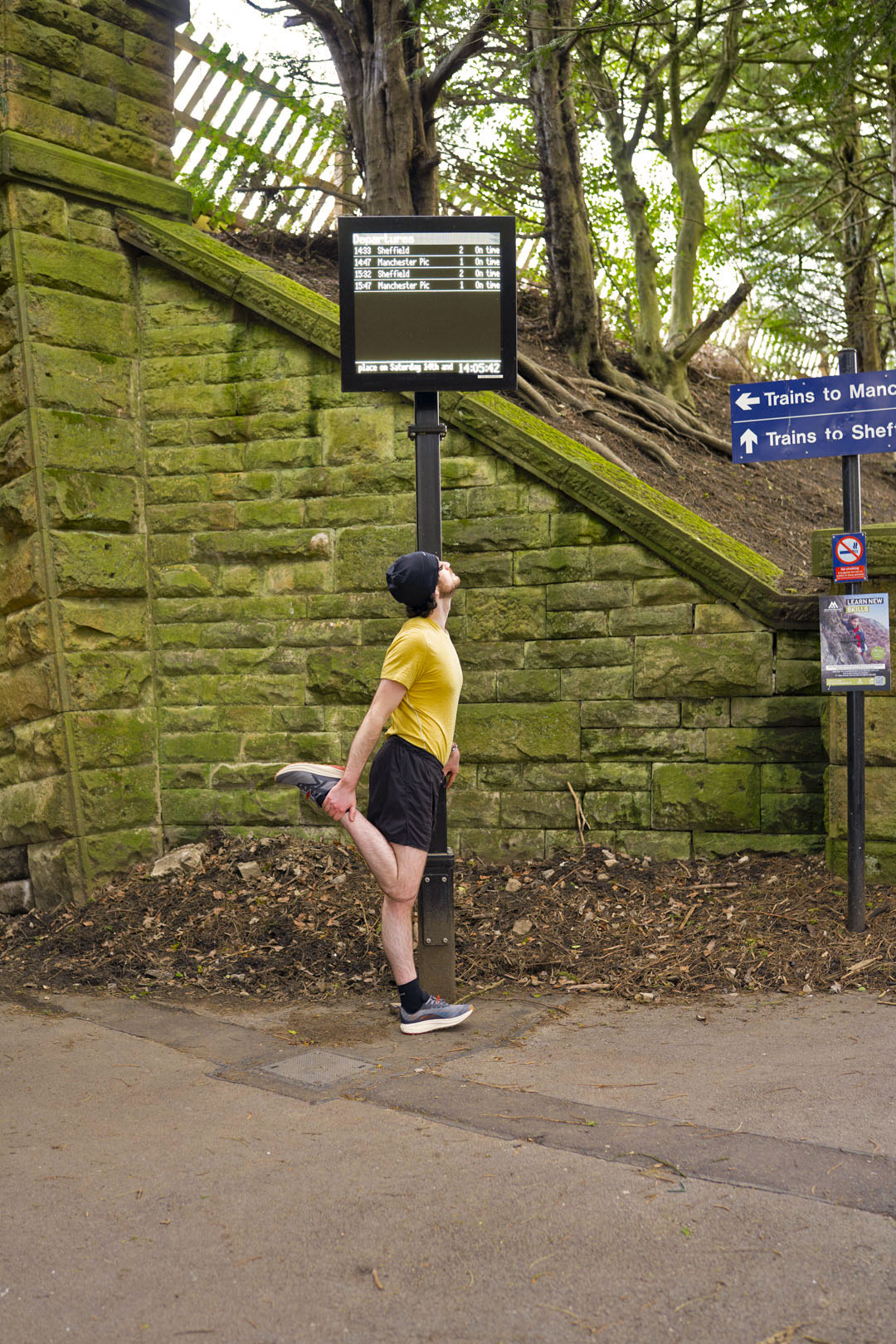 Person stretching outdoors near a stone wall wearing Keen Roam trail running shoes in black/grey colour with signs and trees in the background