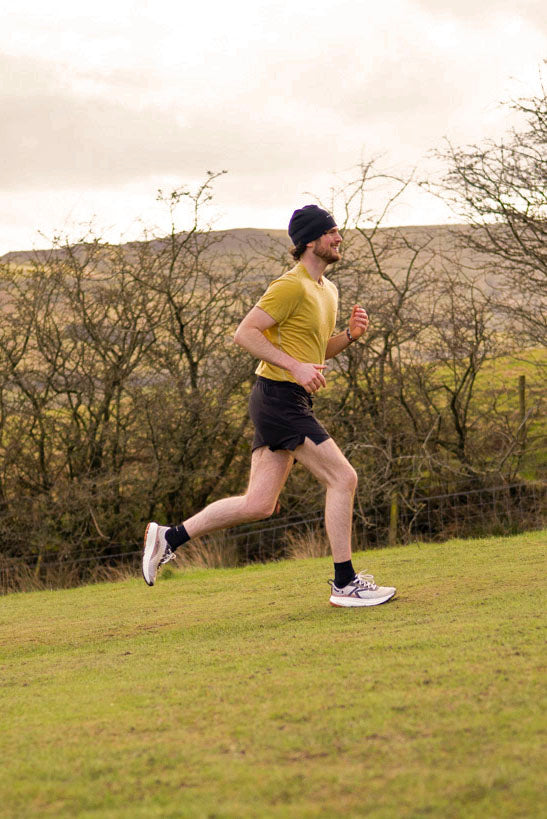 Man wearing a yellow t-shirt running on a grassy field with trees and hills in the background