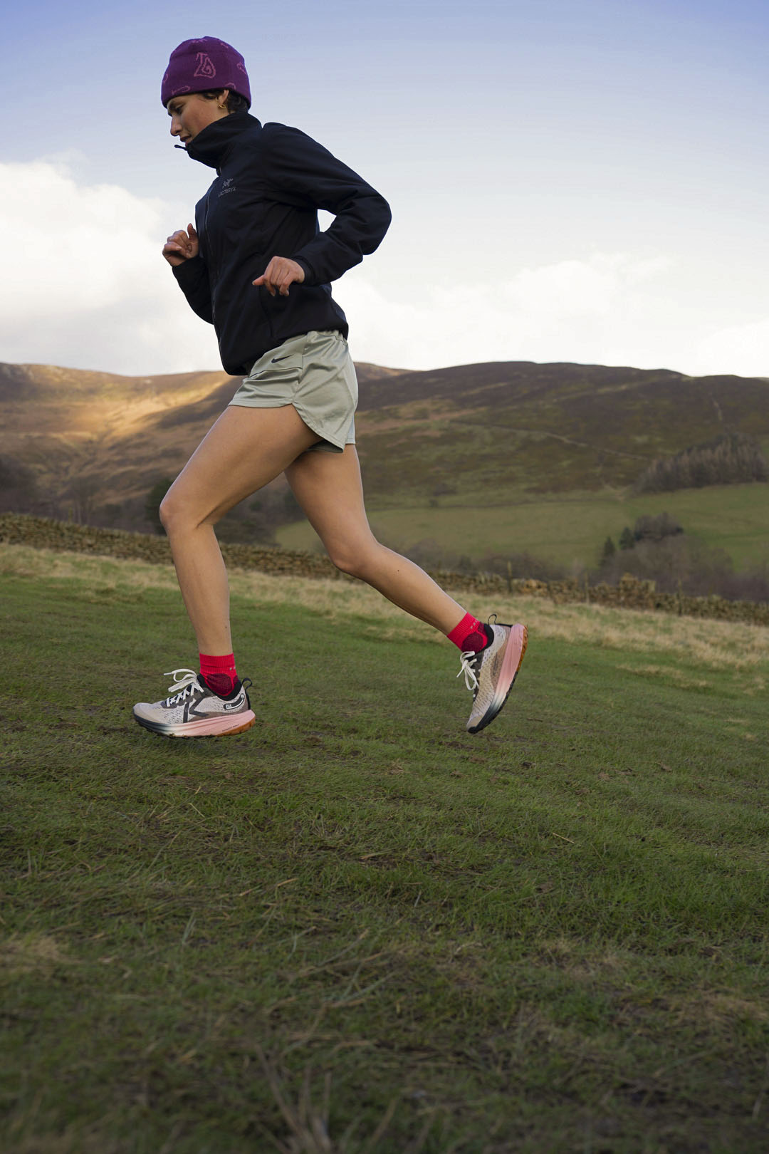 Person running on a grassy field wearing Keen seek trail running shoes with pink socks with hills  in the background