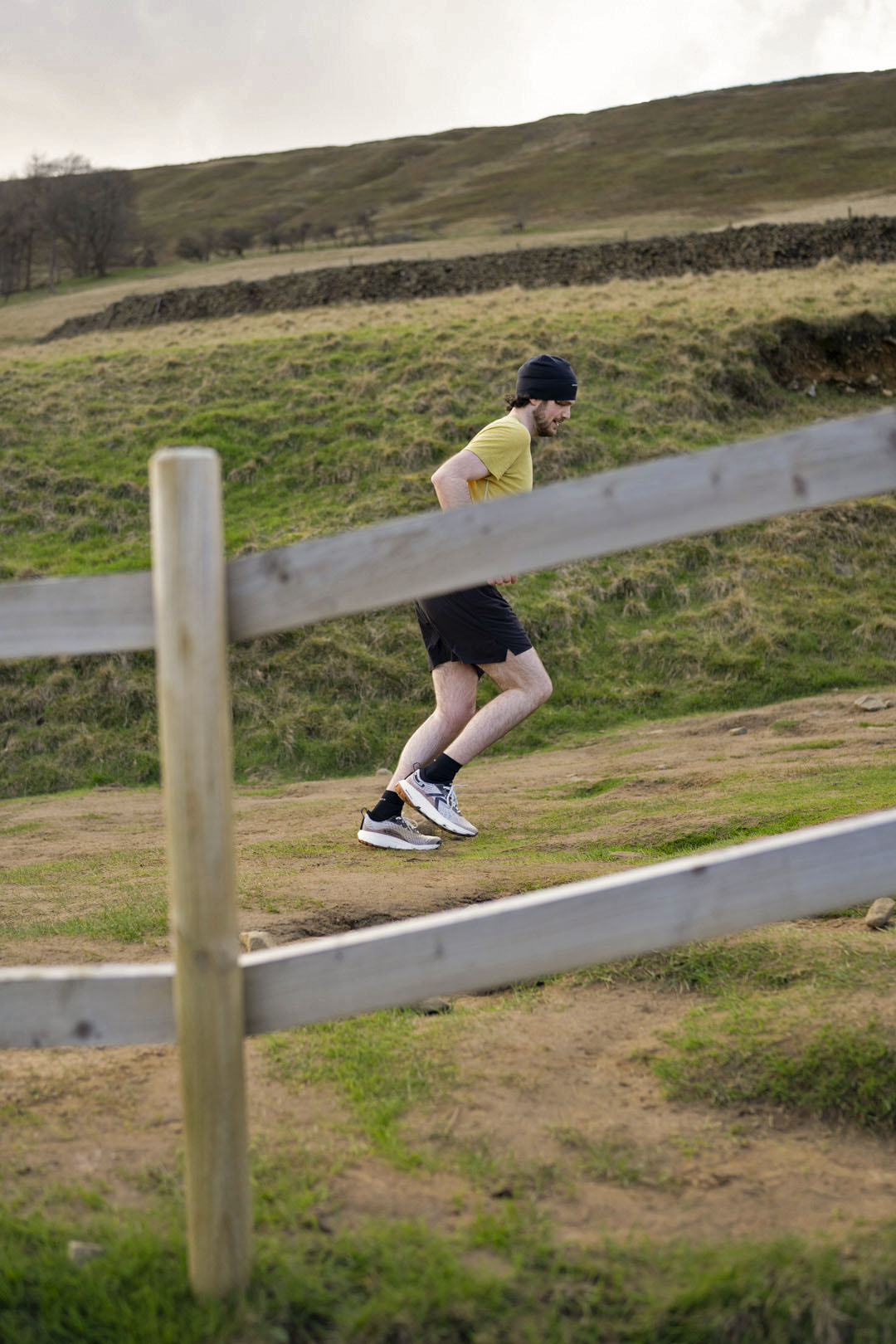 Person wearing a yellow t-shirt and keen trail running shoes running across a wooden fence in a grassy field
