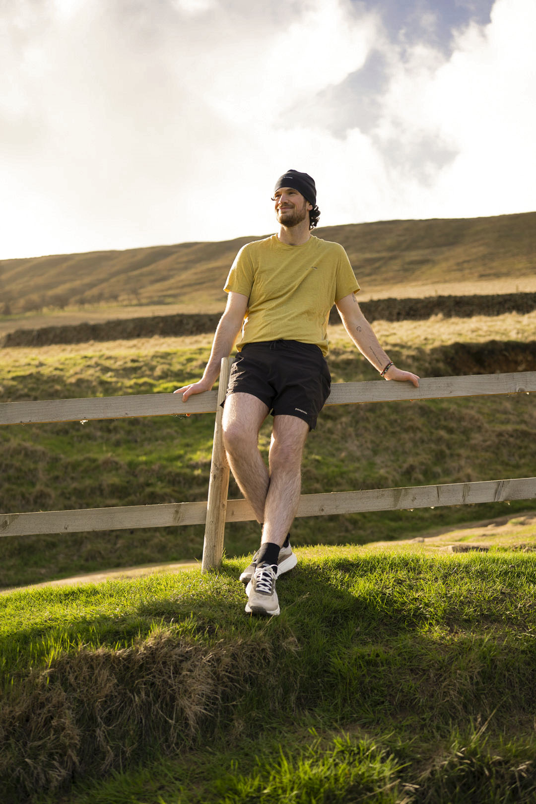 Man in yellow shirt and black shorts standing on a wooden fence in a grassy field with a blue sky.