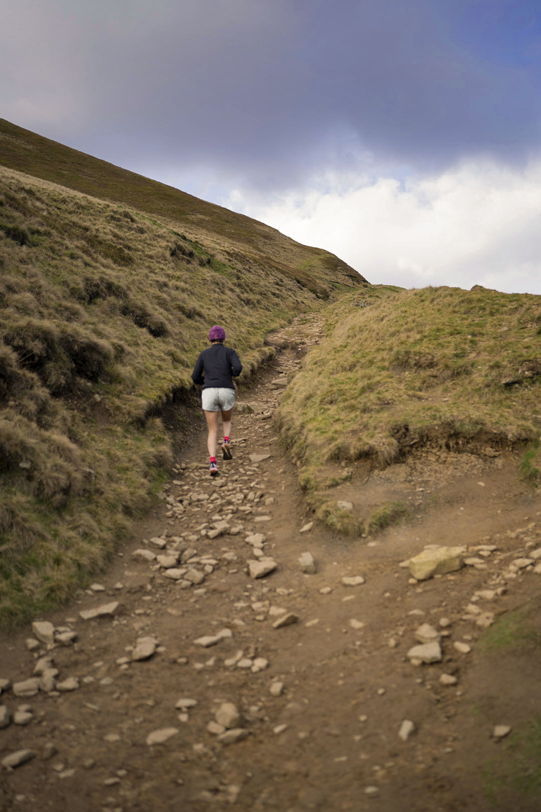 Person hiking on a rocky trail in a mountainous area with grassy slopes and a cloudy sky.