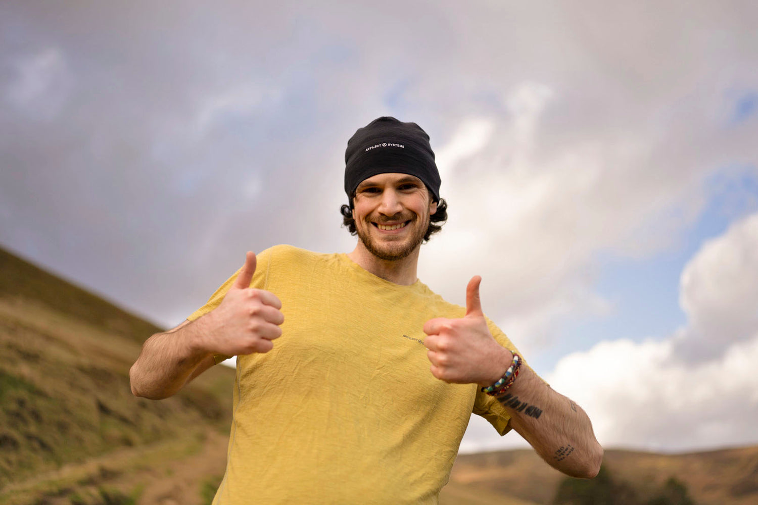 Man in yellow shirt and black cap giving thumbs up in a mountainous landscape