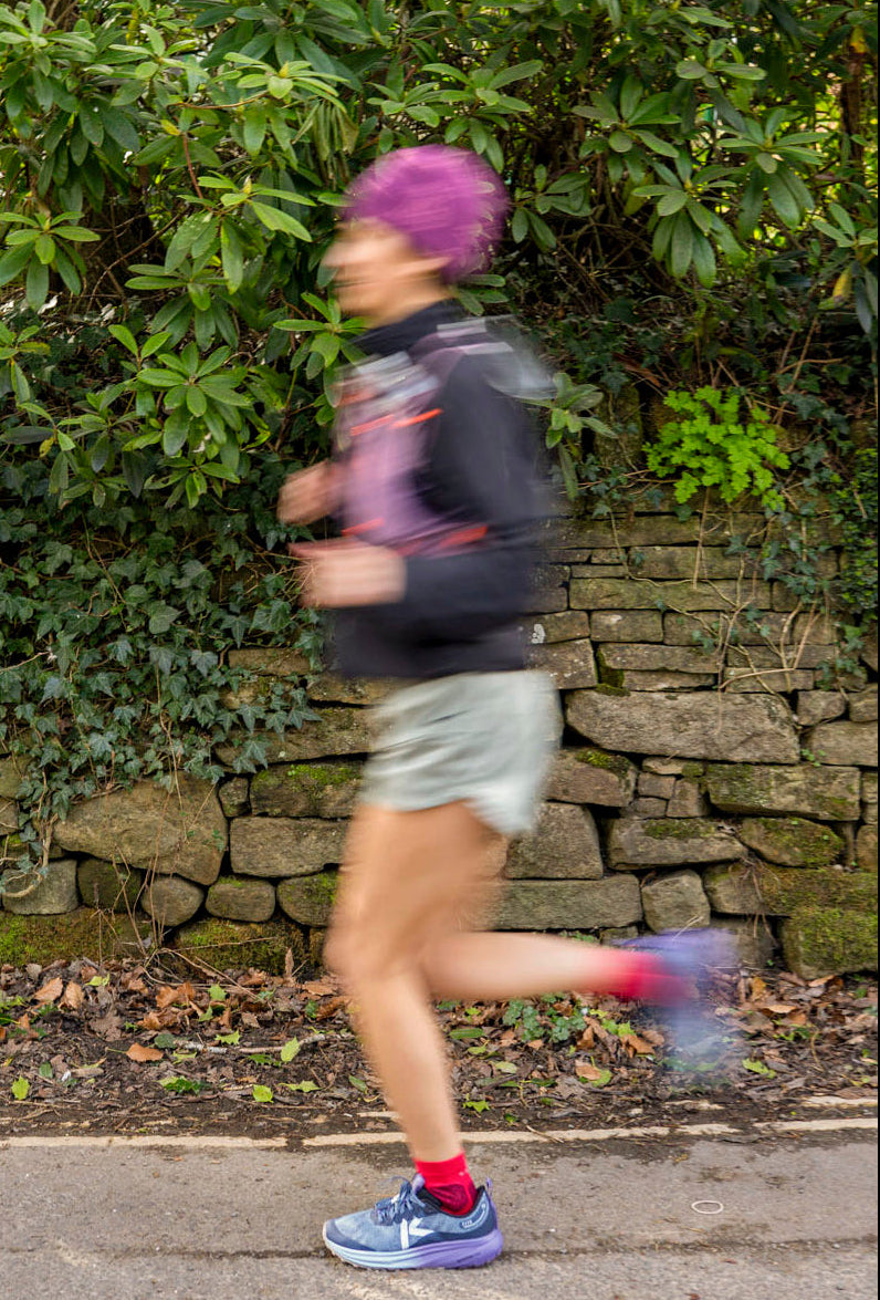 Person running outdoors wearing the keen roam running shoe in blue/purple with greenery and a stone wall in the background