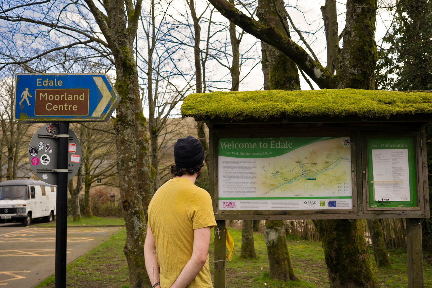 Person looking at an information board in a park with a sign for Edale Moorland Centre wearing a yellow t-shirt and a black hat 