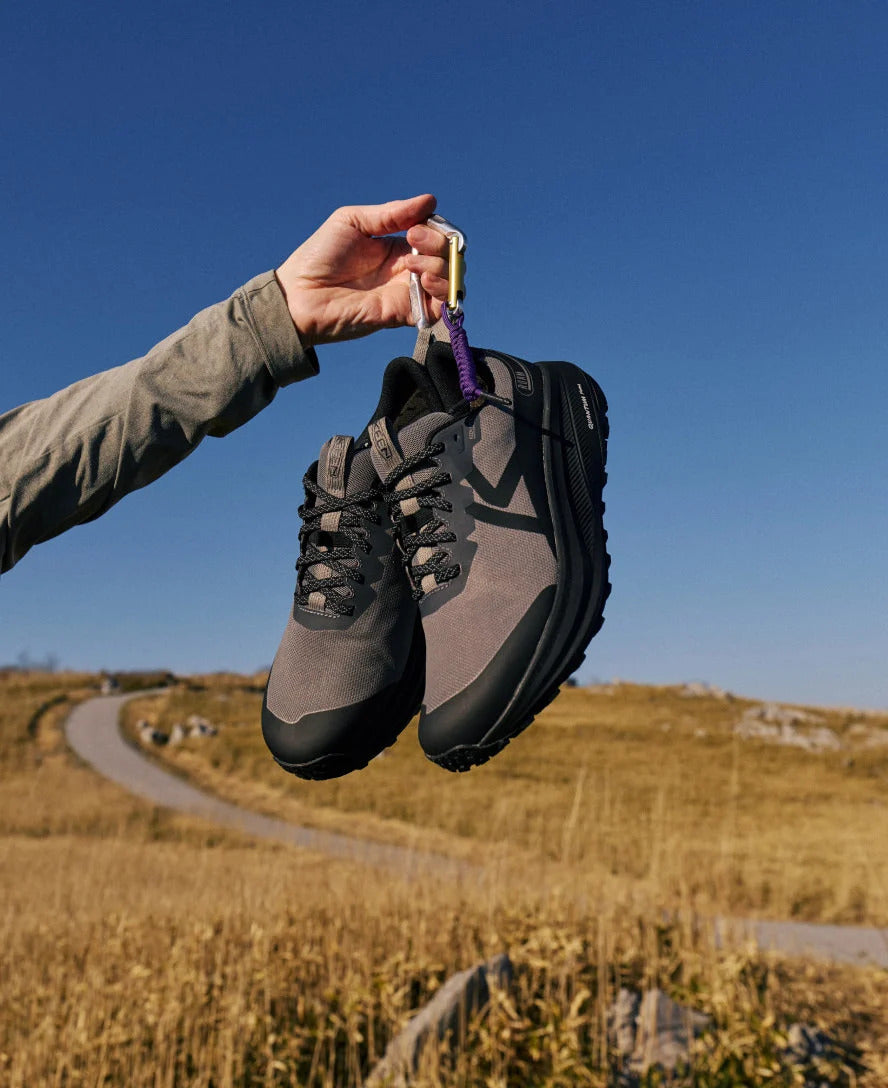 Person holding a pair of hiking shoes with a keychain against a blue sky and path background