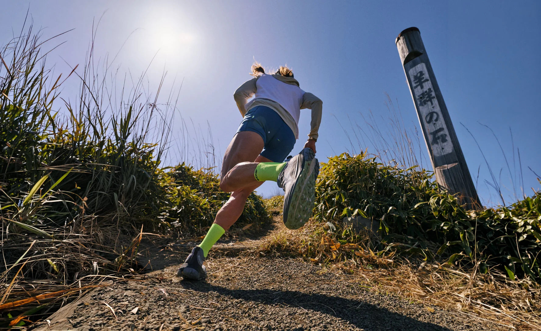 A trail runner in athletic wear sprints up a dirt path outdoors under a bright blue sky, with tall grasses on both sides and a metal signpost with Japanese text to the right.