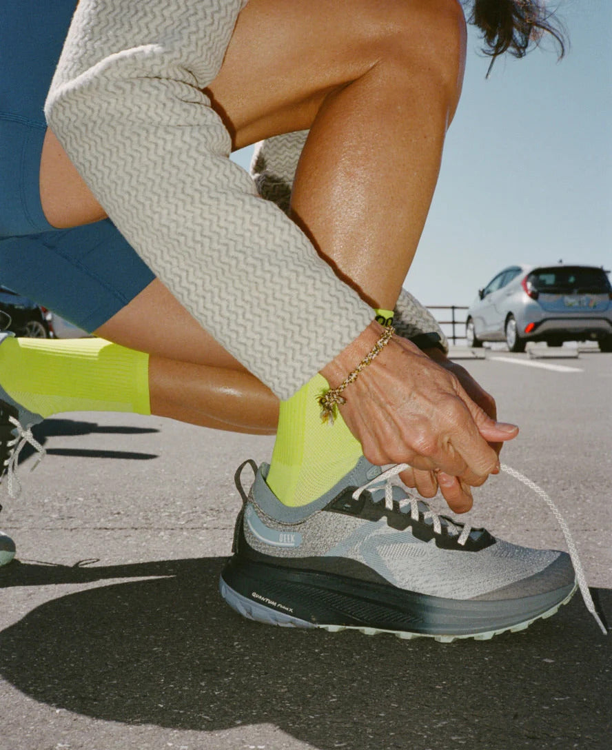 A close-up lifestyle shot of a runner tying the laces of a gray and black trail running shoe, wearing a light beige zigzag knit top, blue shorts, and neon yellow socks in an outdoor parking lot.