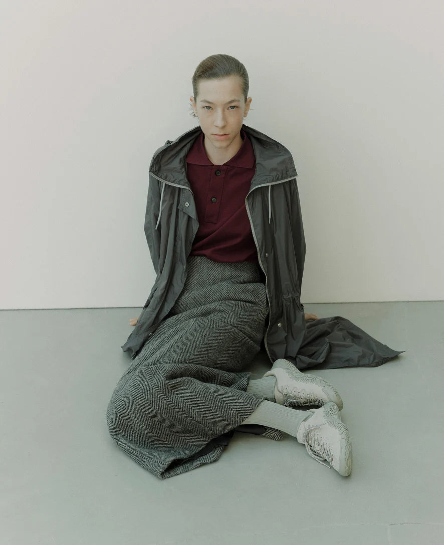 A model sits on a gray floor against a light gray wall, wearing a dark gray hooded jacket