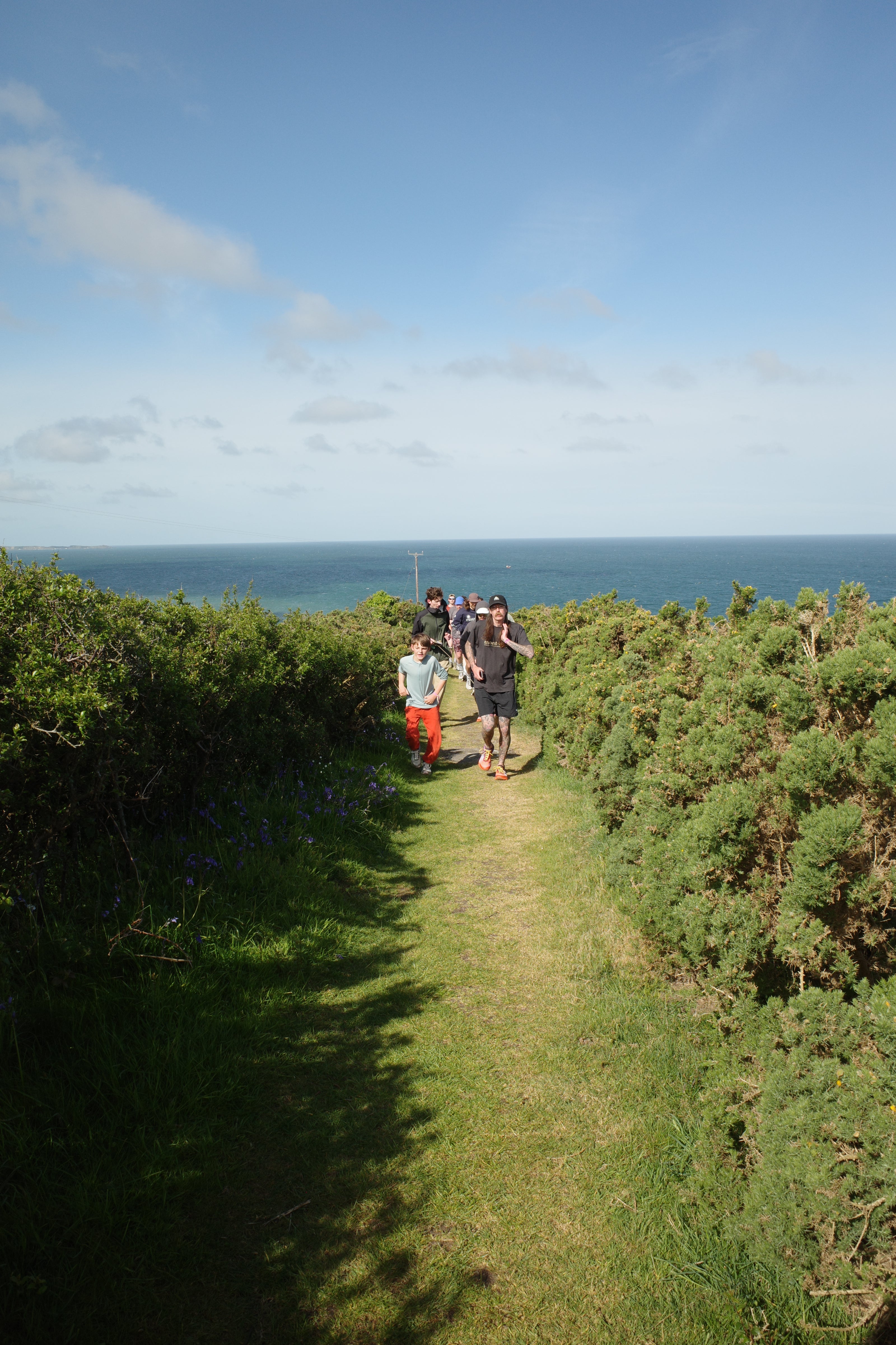 People walking along a path between green hedges with a blue sky and ocean in the background
