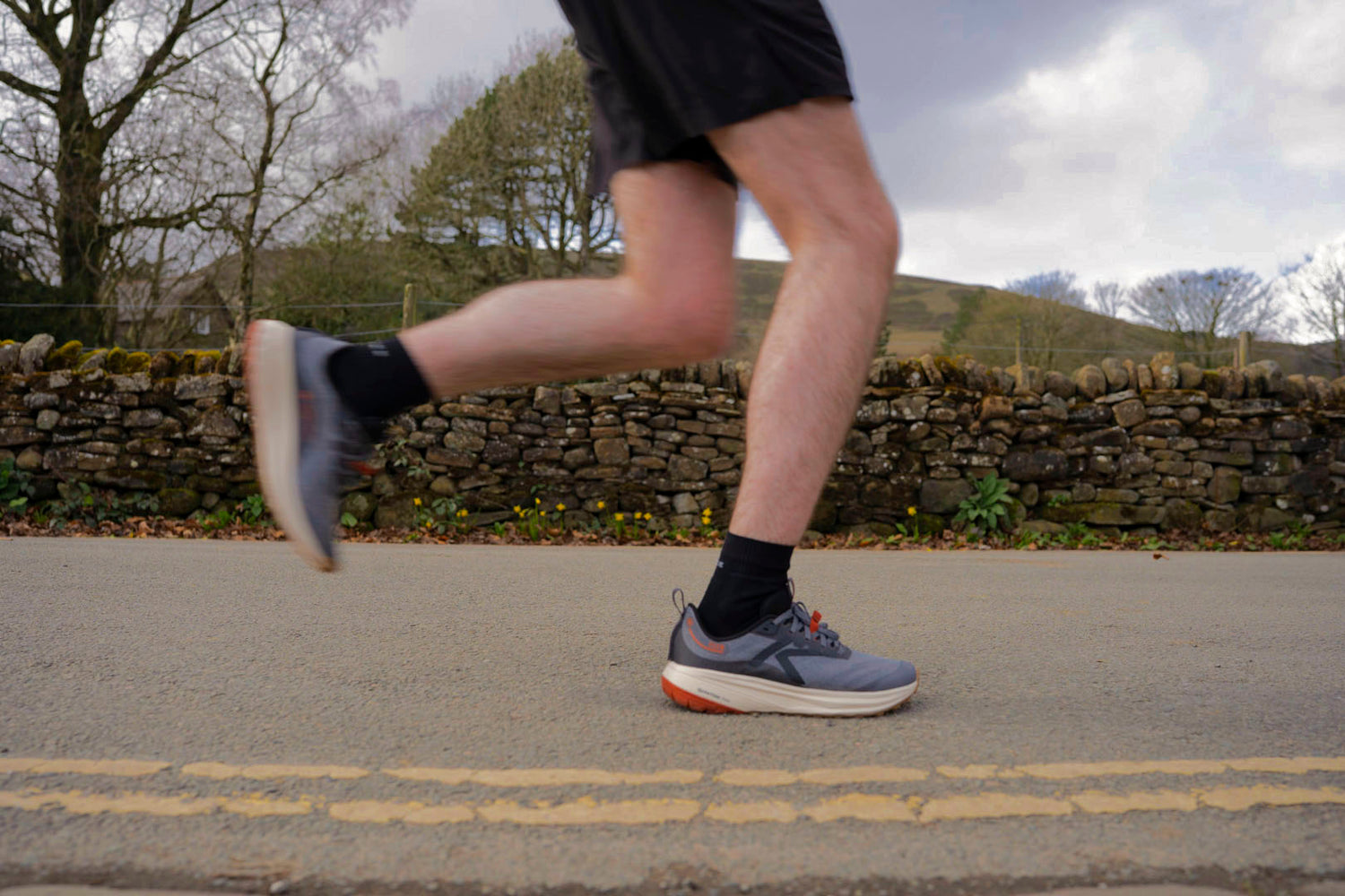 Person running on a road with a stone wall and trees in the background wearing Roam Trail Running shoes 