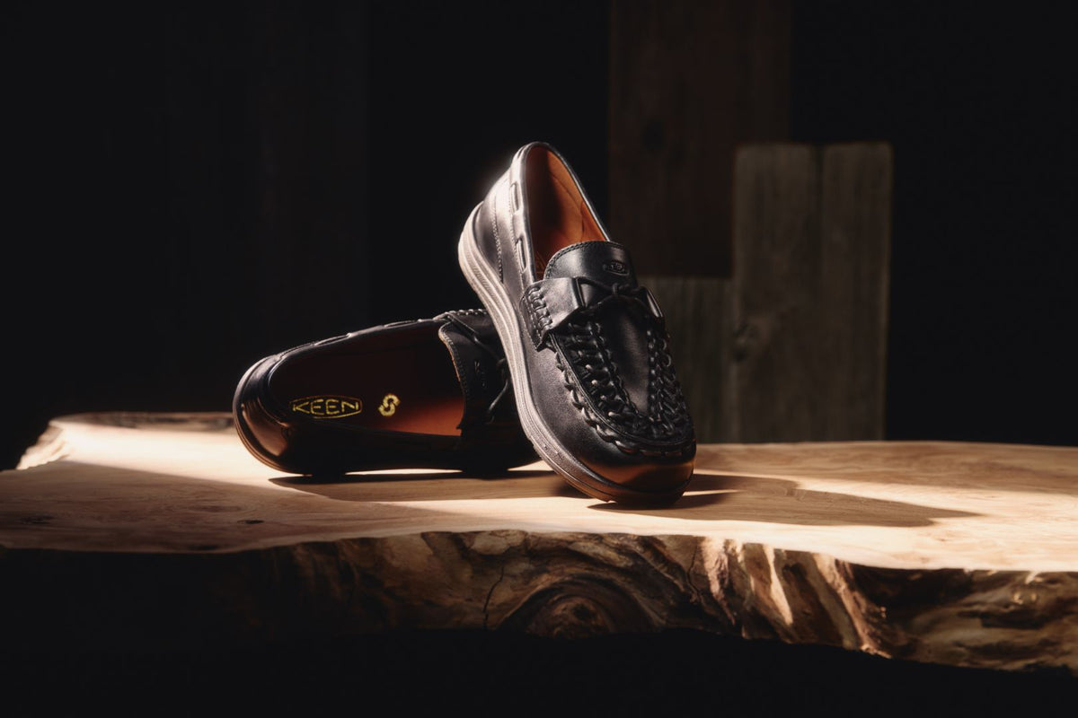 A pair of black leather loafers with braided detailing on the sides, displayed on a rustic
