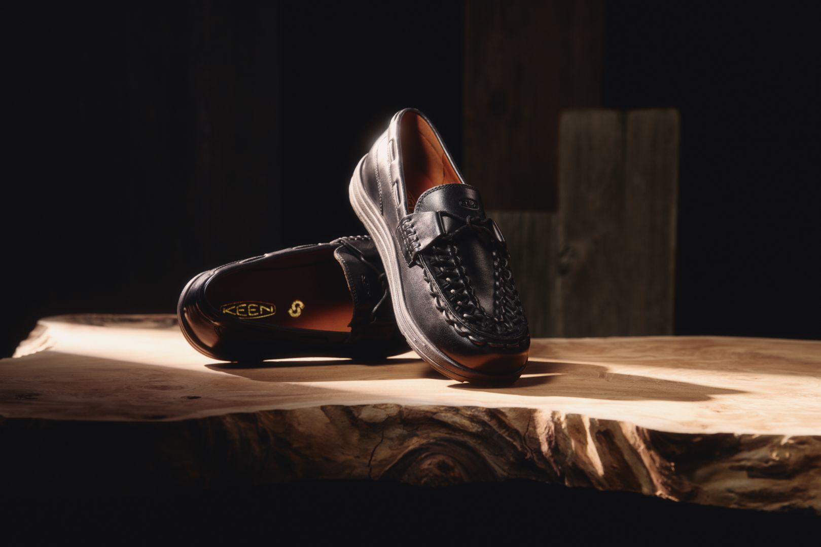 A pair of black leather loafers with braided detailing on the sides, displayed on a rustic live-edge wooden table against a dark background. One loafer is standing upright while the other lies on its side, showing the interior and a gold-toned KEEN logo.