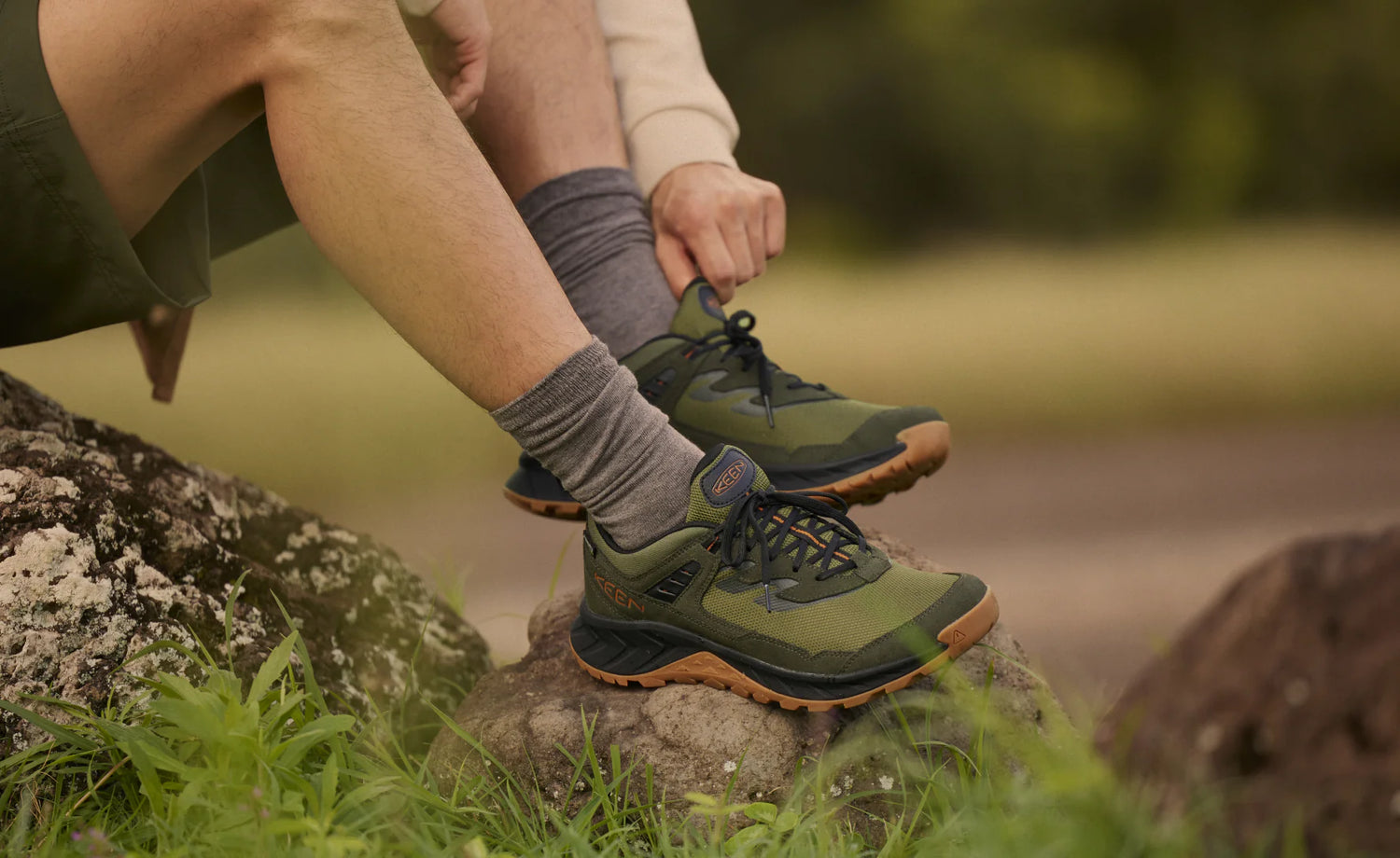 A man wearing the keen hightrail hiking shoes surround by rocks, grass, and trees.