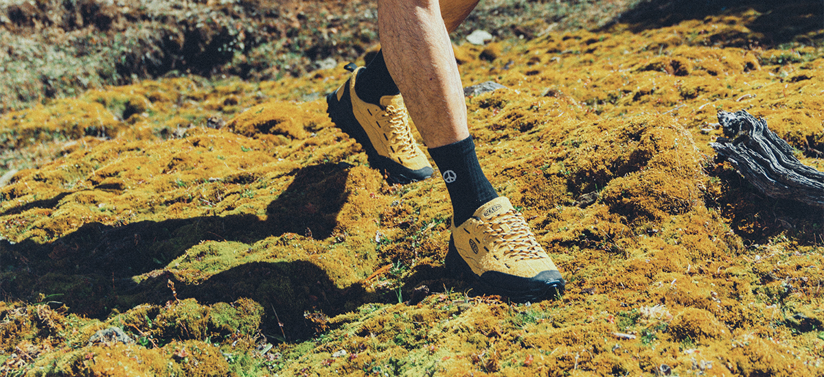 A close-up lifestyle shot of a person's legs walking on a mossy, outdoor trail, wearing tan and black trail running shoes and black socks.