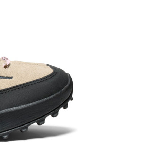 Close-up of a shoe showing the side profile with a beige suede-like upper, black trim, and a black sole with small protruding studs, photographed on a white background.