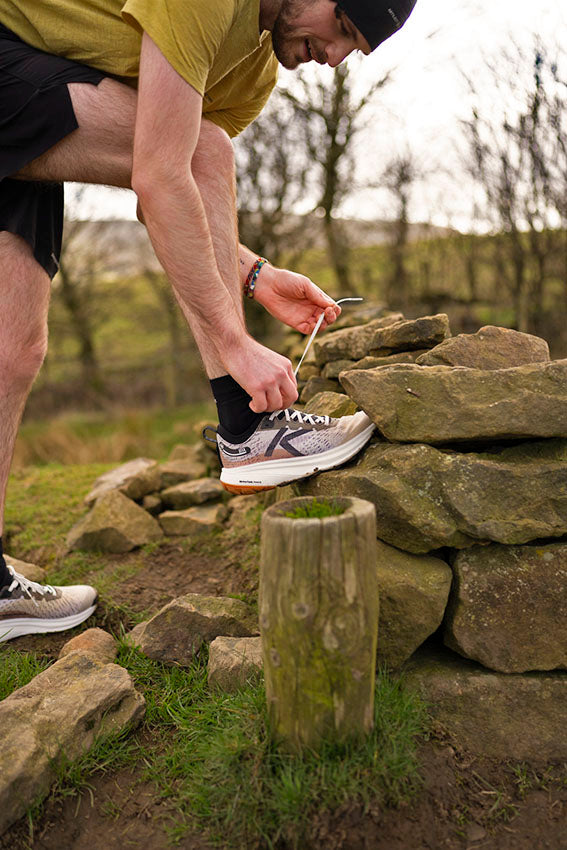 Person tying their shoelace wearing keen trail running shoes with their foot lent on a rock wall.