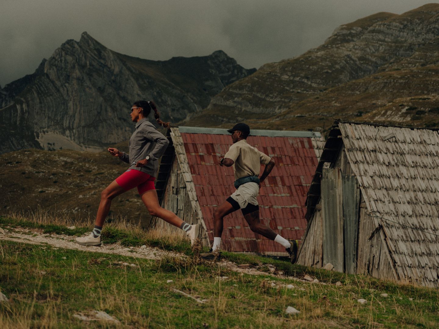 Two people running on a trail with mountainous landscape and rustic buildings.
